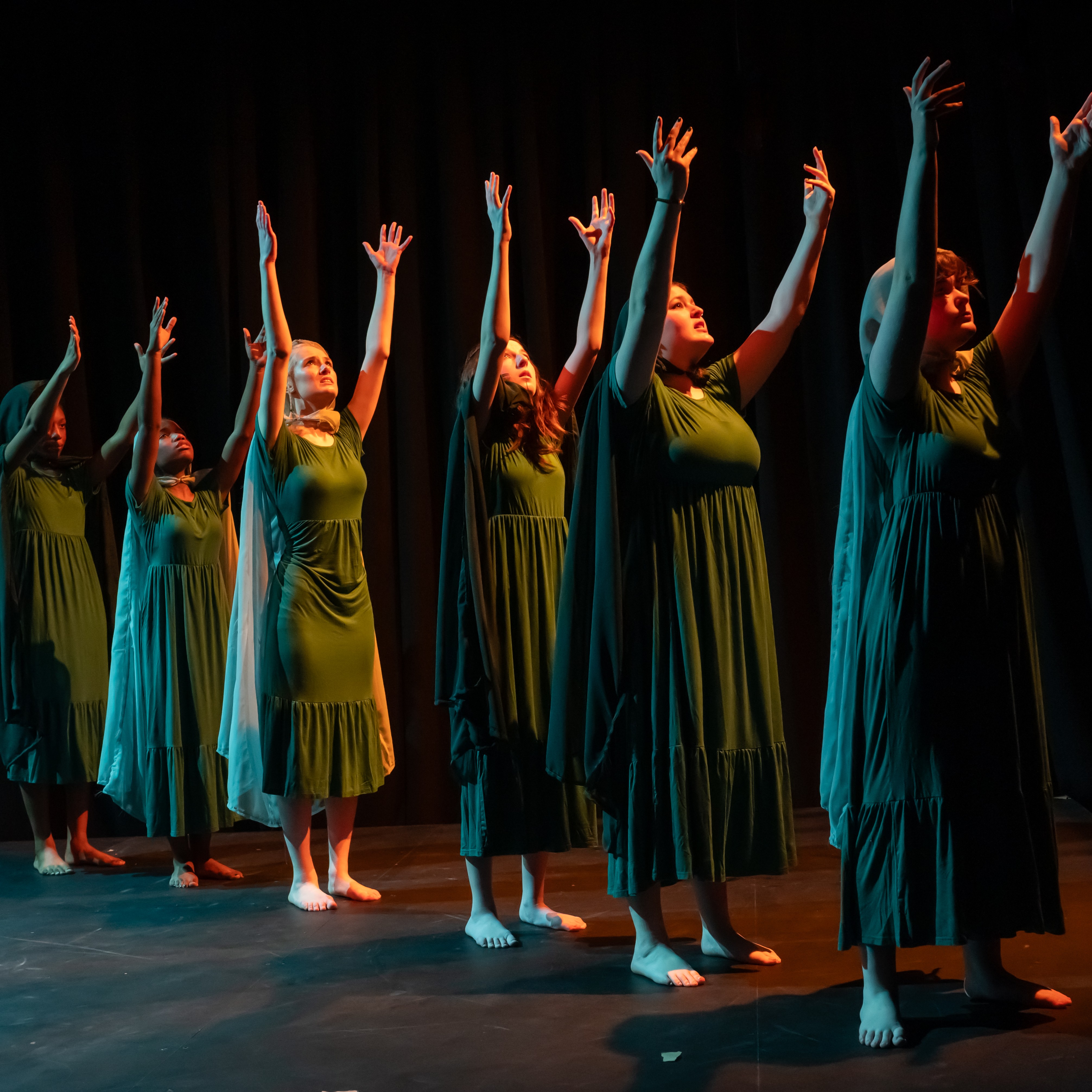 Women in green robes stand in a diagonal line from the back left to the front right. Their arms are raised and they are surrounded by amber light.